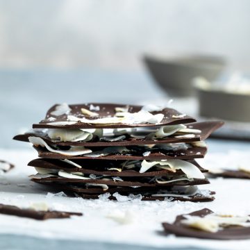 side view of a stack of thin chocolate bark to show the thickness of the chocolate. Coconut flakes and sea salt sprinkled around the foreground and background