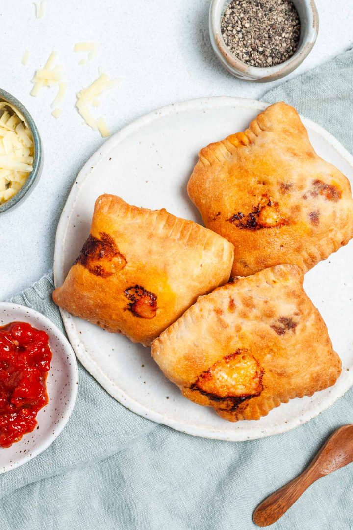 overhead photo of three golden brown cooked pizza roll pockets on a plate, the backdrop sprinkled with grated cheese
