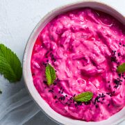 overhead photo of beetroot raita in a bowl on white background, decorated with fresh mint leaves and nigella seeds