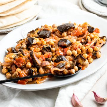 A white serving plate of Maghmour with a serving spoon ready to serve. A plate piled with flatbread just visible in the background.