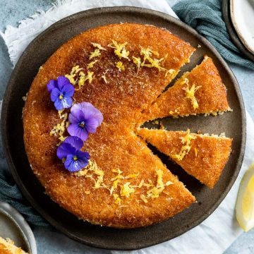 a baked lemon syrup cake on blue plate with three pieces cut out, one on a plate. The cake is decorated with a ring of lemon zest and purple edible flowers