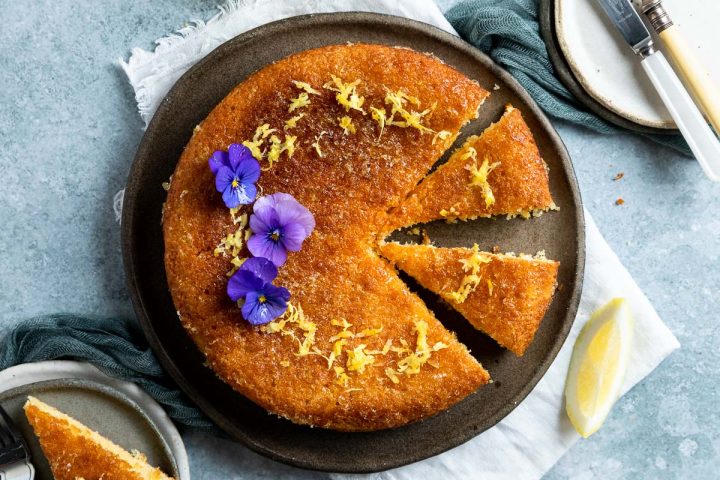 a baked lemon syrup cake on blue plate with three pieces cut out, one on a plate. The cake is decorated with a ring of lemon zest and purple edible flowers