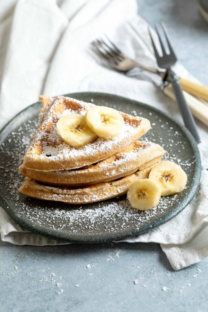 a stack of three sections of waffles sprinkled with icing sugar and topped with sliced banana