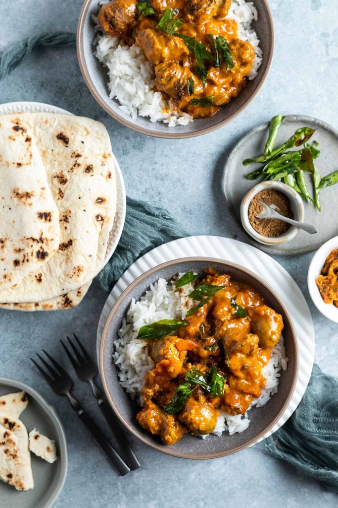 two bowls of curry served with rice, naan bread, fried curry leaves and chutney on the side