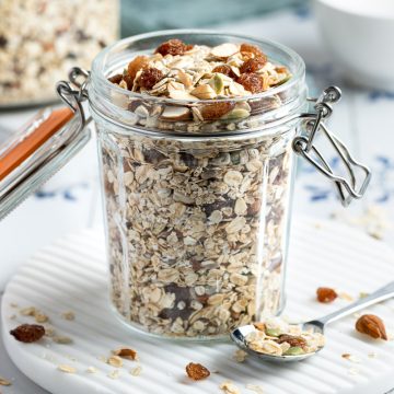 muesli in glas storage jar, a spoon of muesli to the right of the jar with oats, nuts and fruit spilled over the table.