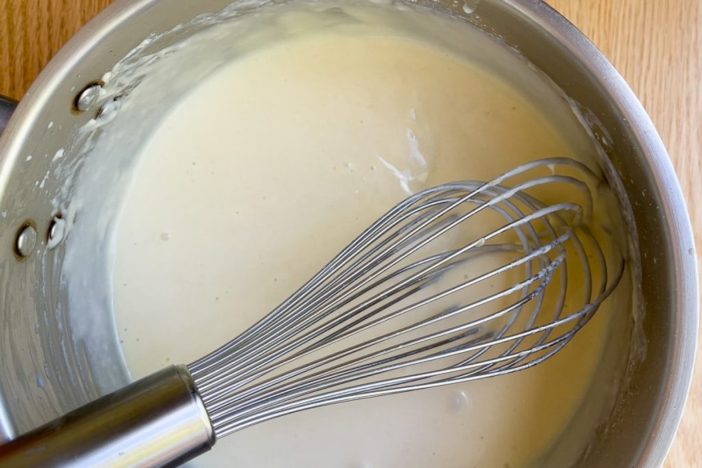 whisk in pan with sauce to show the consistency of sauce with half the milk added.