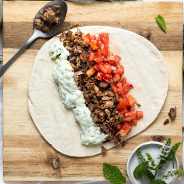 Pita bread on board topped with cooked lamb, tomato salad and tzatziki ready to roll up and make Greek lamb wrap