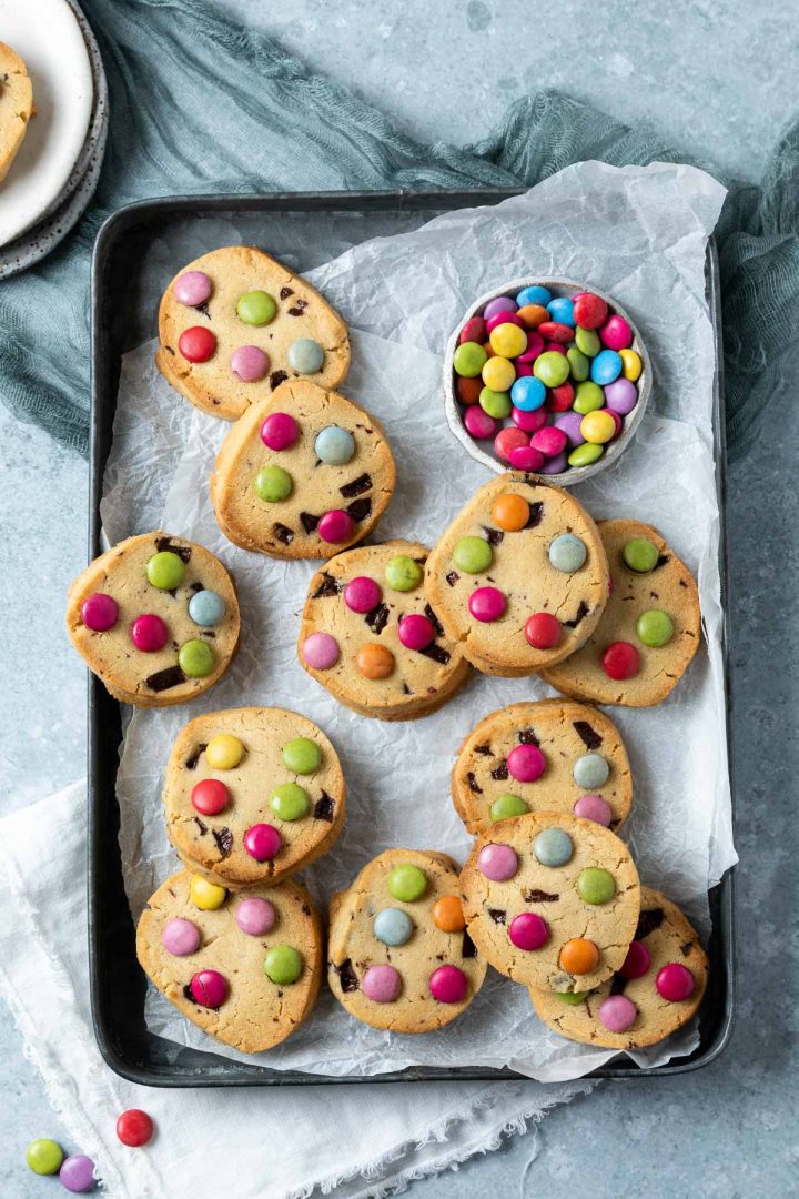 a batch of Smartie cookies on a tray with a small dish of Smarties in the top right.