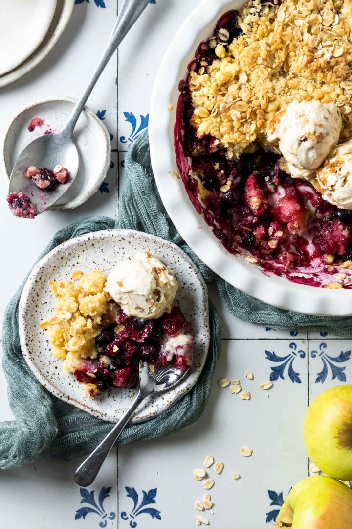 apple and blueberry crumble served on a plate with a scoop of ice cream, the rest of the crumble in the dish just visible to the top right,