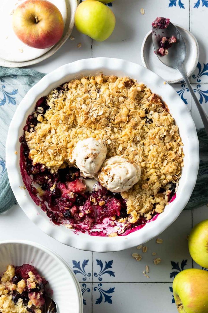 apple and blueberry crumble in baking dish topped with two scoops of ice cream, a few fresh apples just visible at the edges of the image.