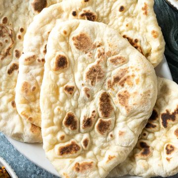 fresh homemade naan bread on a white plate, with a bowl of curry just visible in bottom left corner.