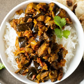 Aubergine curry served in a white bowl with rice, naan bread on the right hand side of the bowl.