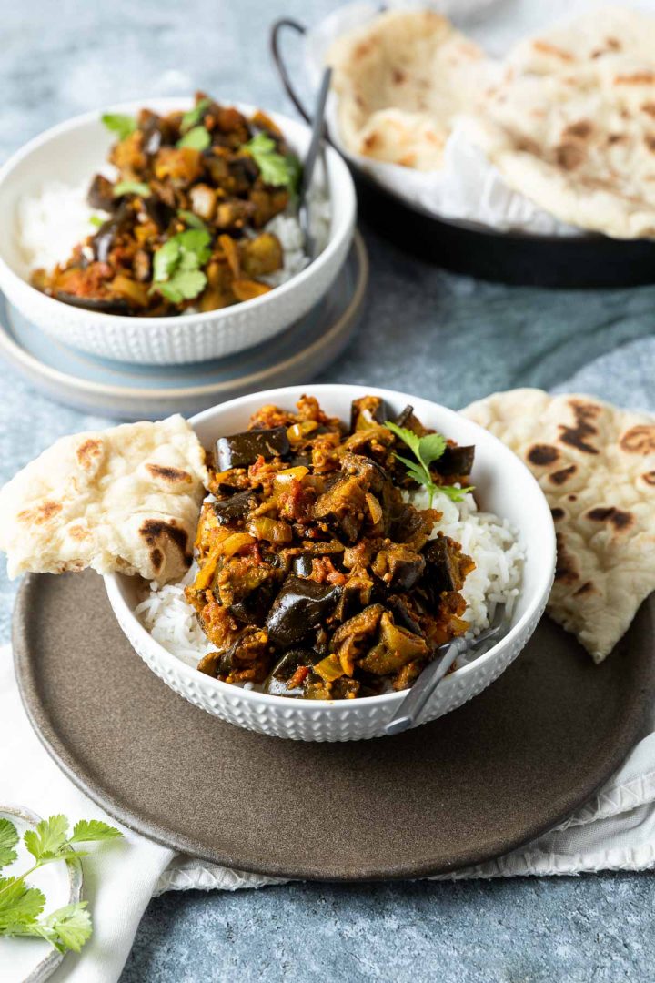 two bowls of aubergine curry served with rice, with a plate of naan bread just visible at the back of the photo.