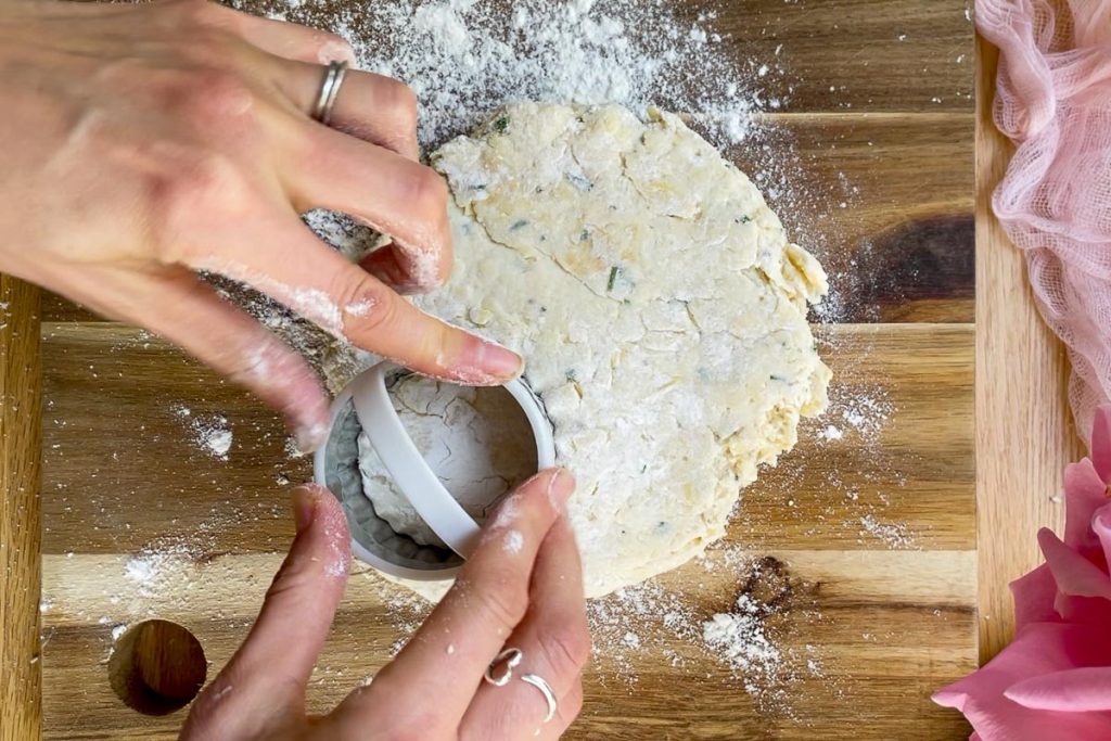 The rolled scone dough on a floured wooden board with a scone cutter being pressed in to the dough.