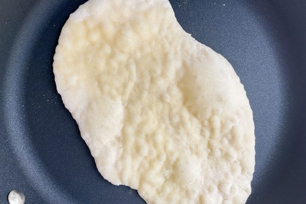 A naan bread in a frying pan with large bubbles in the dough.