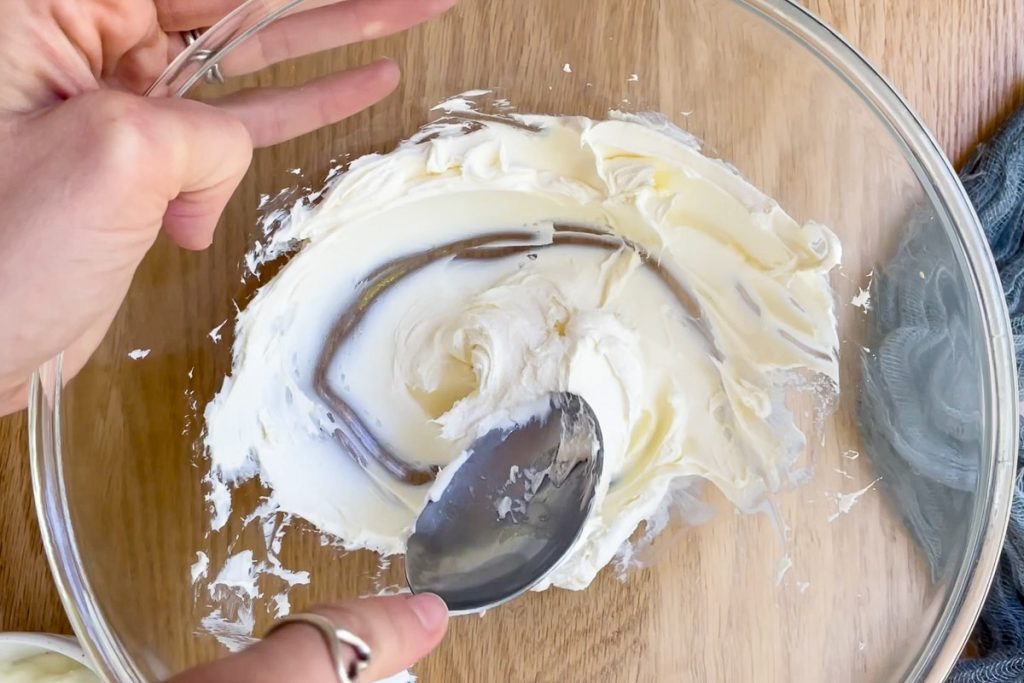 Cream cheese being mixed with a spoon in a mixing bowl to soften it.