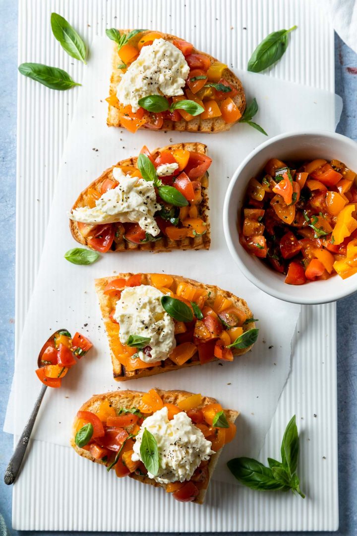 Bruschetta on a white board with a small bowl of chopped tomatoes and a spoon.