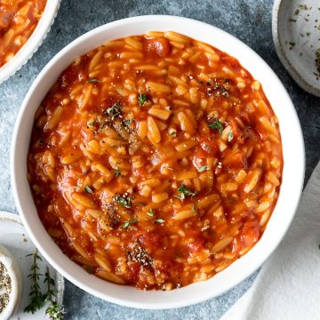 A white bowl of Manestra, Greek tomato orzo, with extra Greek oregano on the side to sprinkle over.