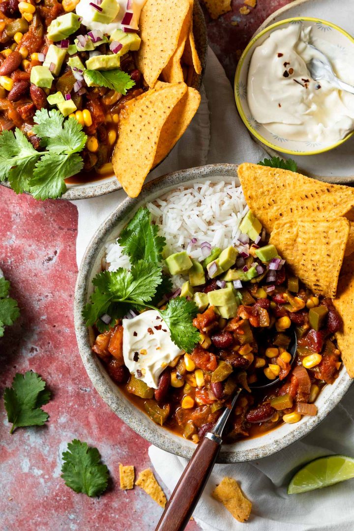 Two bowls of bean chilli served with rice and corn chips, with a bowl of sour cream to the top right corner.