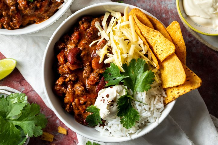 slow cooker chilli con carne in a bowl with rice and corn chips, topped with grated cheese, sour cream fresh corainder.