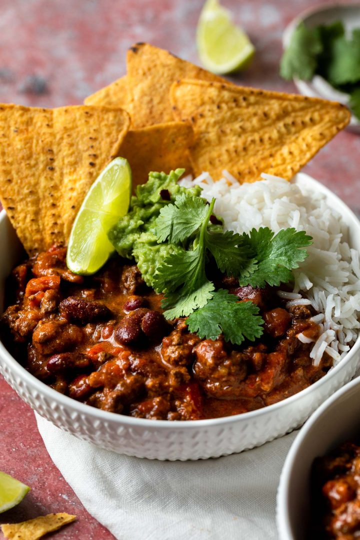 A bowl of slow cooker chilli served with corn chips and rice.