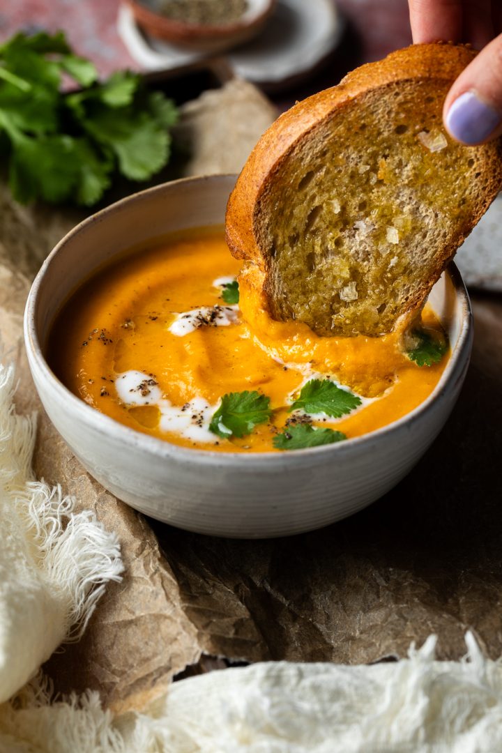 A hand dipping a slice of toasted bread into the bowl of carrot soup.