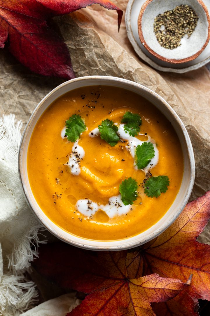 A bowl of carrot soup topped with coconut milk, with red Autumn leaves in the background.
