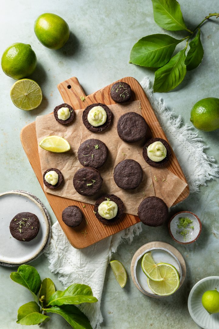 Chocolate lime cookies on a wooden board, some sandwiched with lime buttercream, others topped with buttercream.