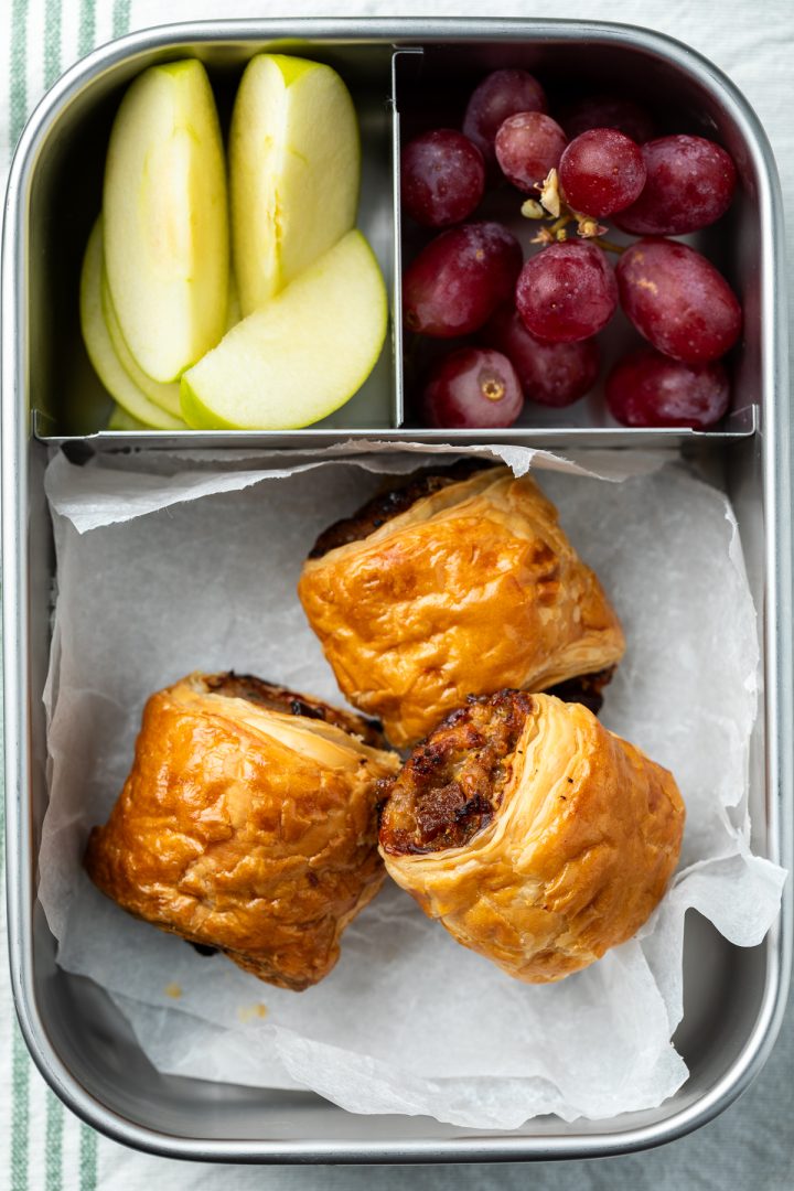 Cheese sausage rolls in a lunchbox with apple and grapes.