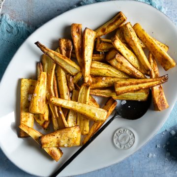 Roast parsnips on a white oval serving plate with extra salt to sprinkle over.