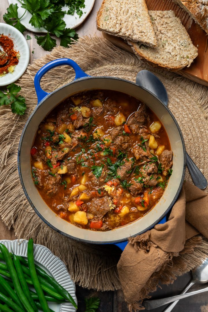 A pot of beef goulash with green beans on the bottom left corner and fresh bread in the top right corner.