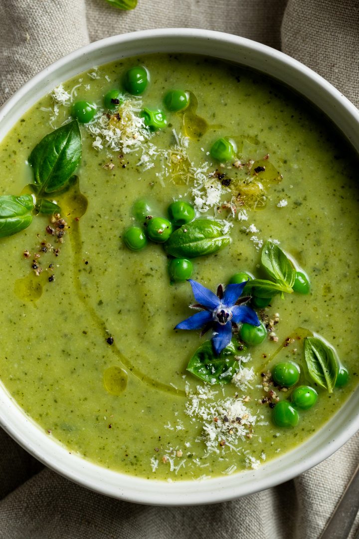 Close up of a bowl of courgette soup (zucchini soup) decorated with peas, parmesan cheese and a blue borage flower.