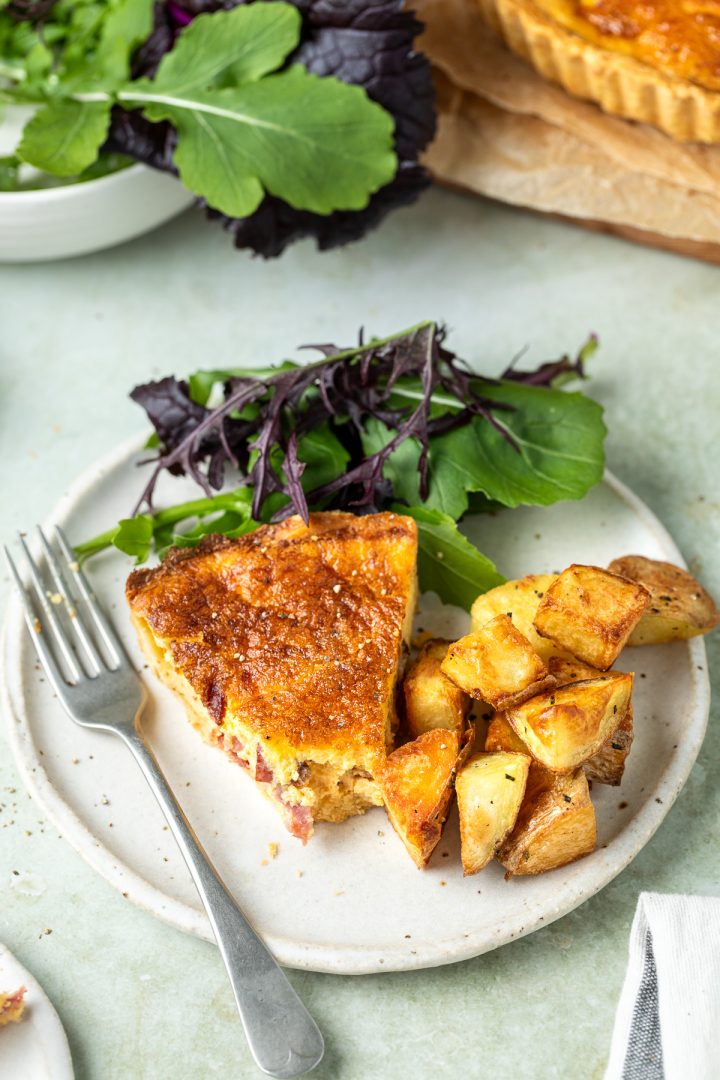 A slice of quiche Lorraine on a plate served with parmentier potatoes and salad leaves.