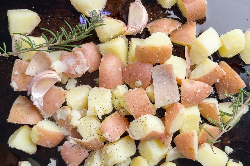 The potatoes on a baking tray with garlic cloves and rosemary sprigs.
