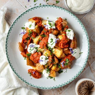 A plate of patatas bravas topped with rosemary flowers, with extra garlic mayonnaise in the top right corner.