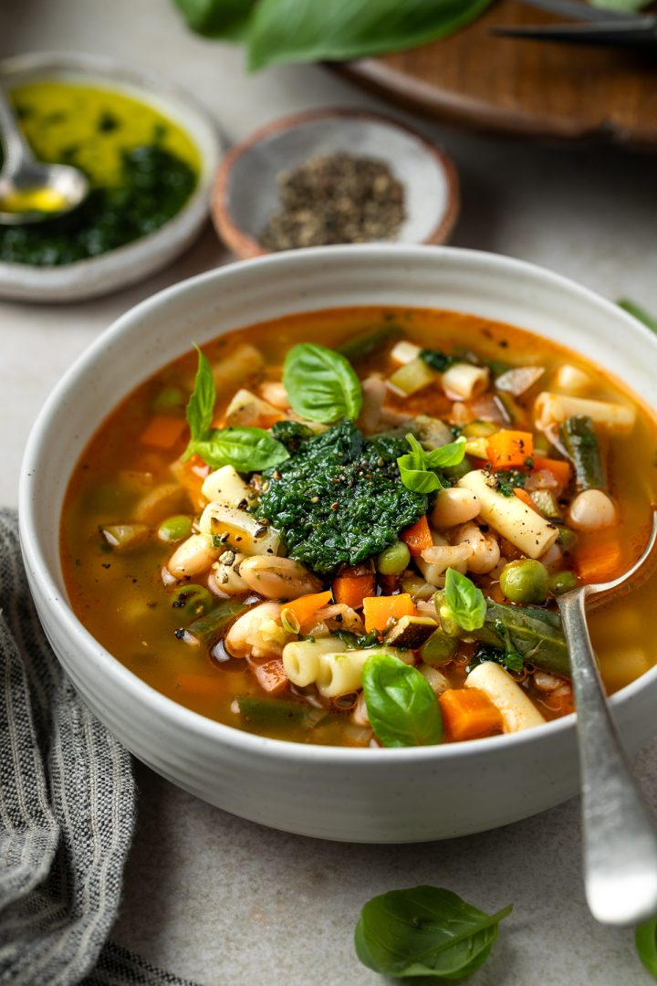 A bowl of vegetable soup with a spoon, extra basil pistou in the background.