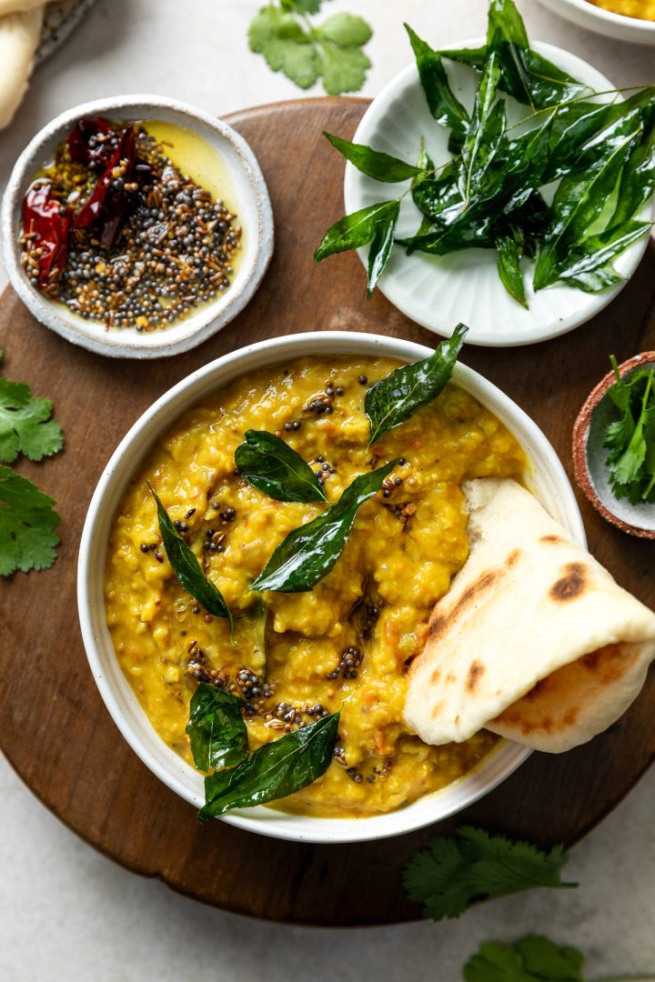 A bowl of red lentil dahl served with a naan bread, with extra fried curry leaves and mustard seeds in bowls to the side.