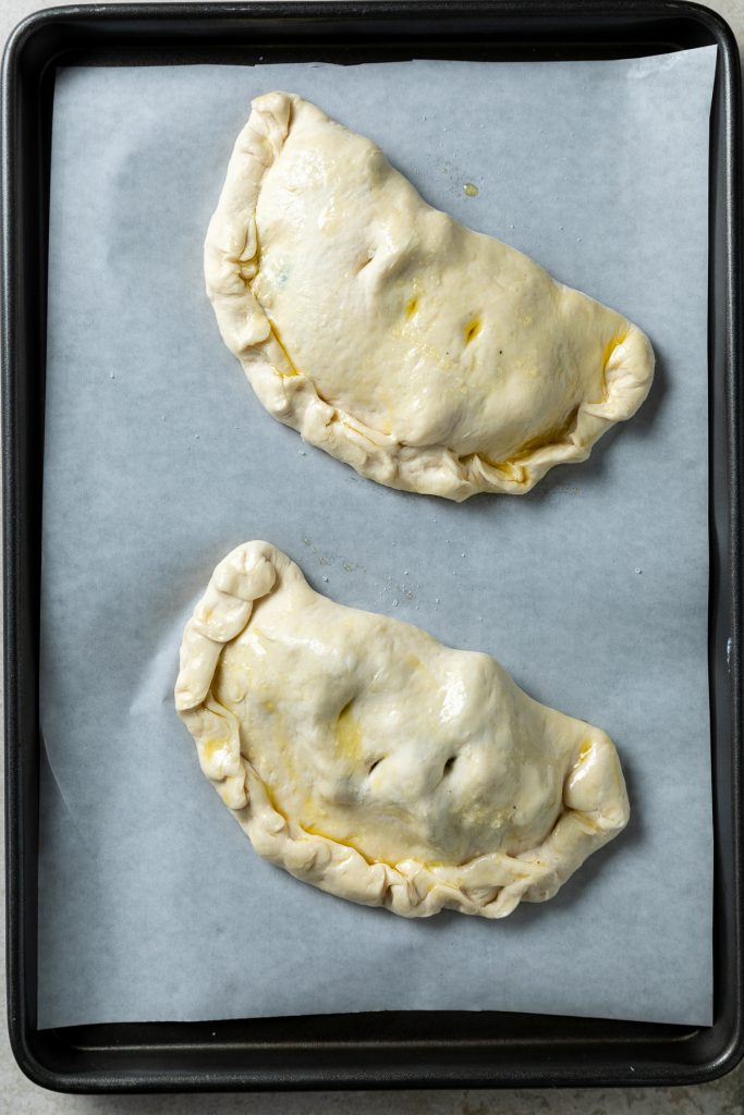 Two unbaked calzone on a lined baking tray.