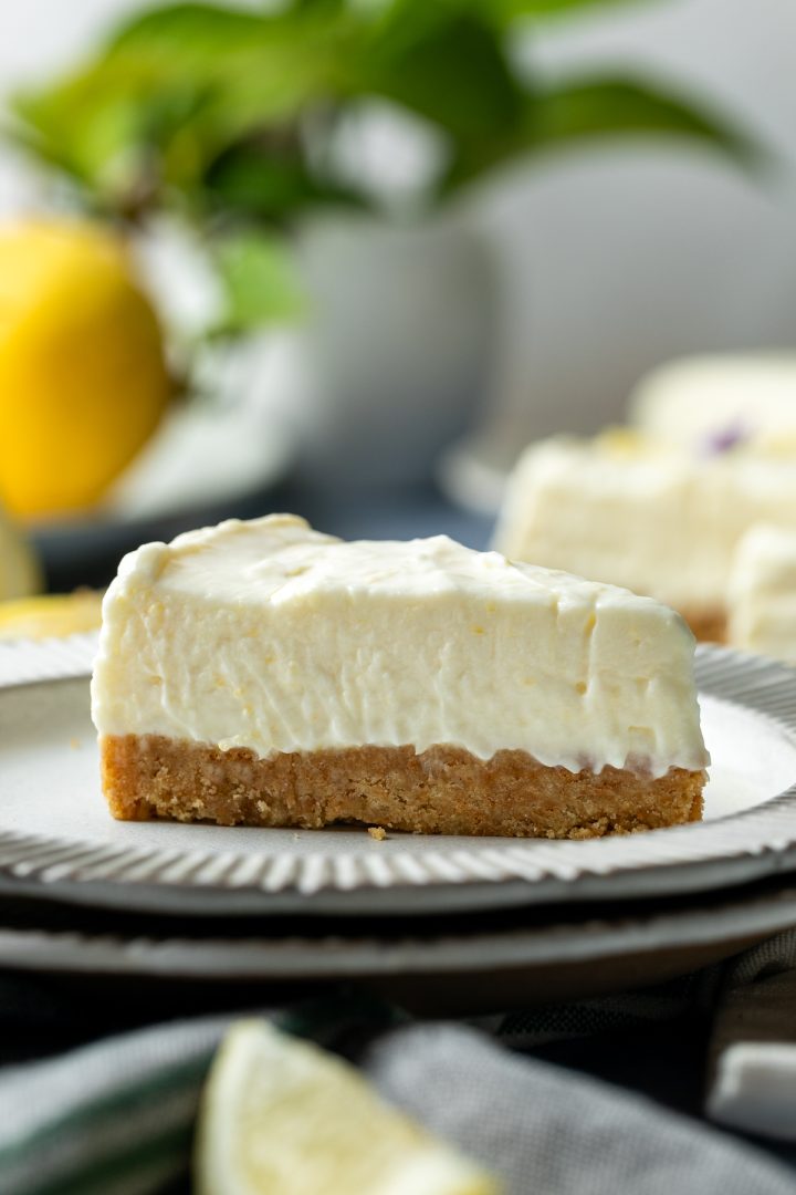 A side view of a slice of cheesecake on a plate to show the biscuit base and creamy set texture of the lemon cheesecake topping.