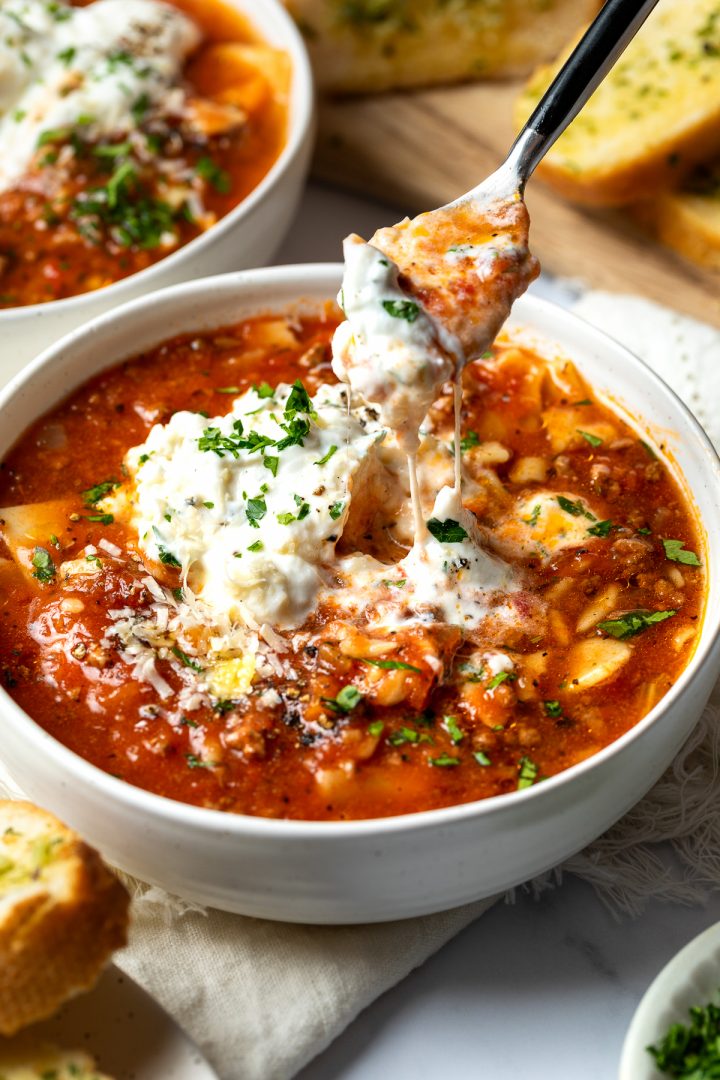 A spoon digging in to the oozing cheese on top of a bowl of lasagna soup.