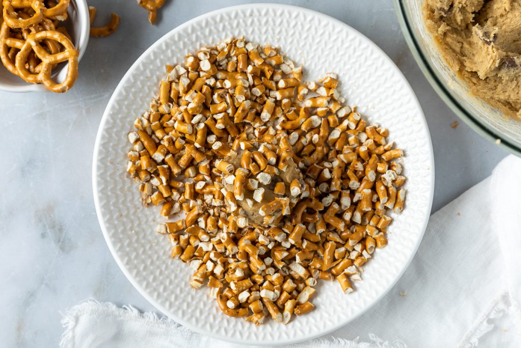 A ball of cookie dough being rolled in the chopped pretzel pieces.