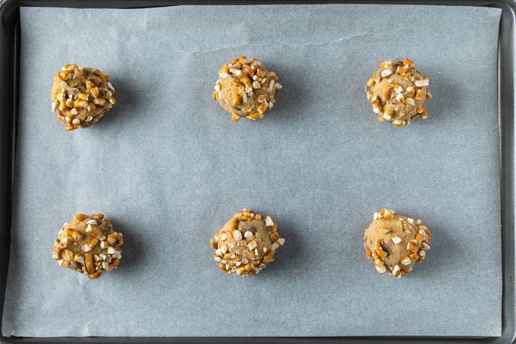 6 cookie dough balls rolled in pretzel pieces on a lined baking tray.