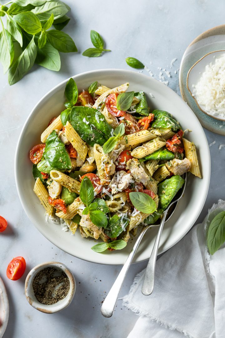 A bowl of chicken and pesto pasta with serving spoons.