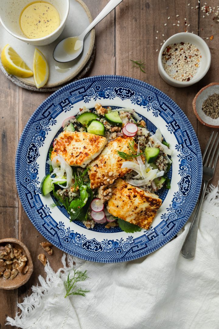 A plate of quinoa salad topped with pan fried halloumi, with extra lemon dressing and walnuts on the side.
