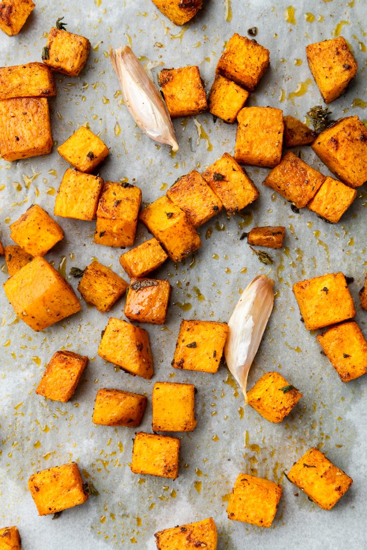 Cubes of roasted butternut squash with whole garlic cloves on a lined baking tray.