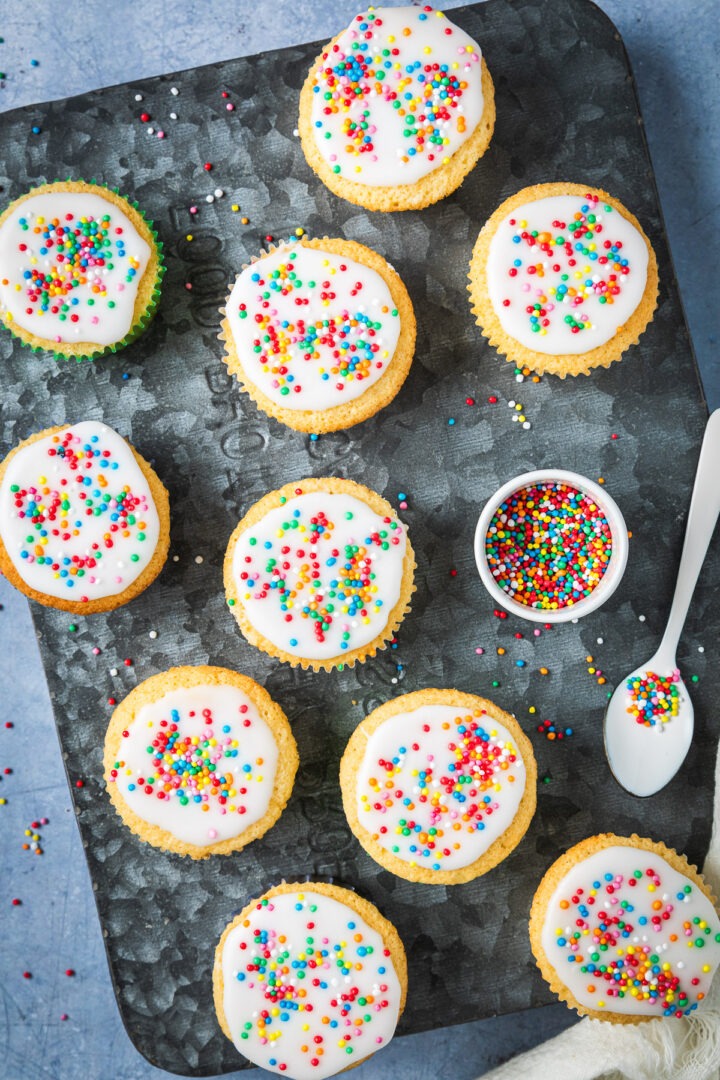 Overhead of freshly iced fairy cakes with a pot of sprinkles.