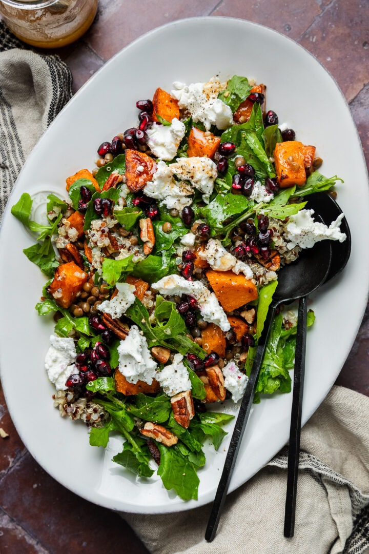 A platter of roast pumpkin quinoa salad topped with pecans and pomegranate seeds with serving spoons.