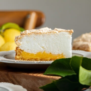 A slice of gluten-free lemon meringue pie on a plate, showing the fluffy meringue, yellow lemon curd and almond crust layers.