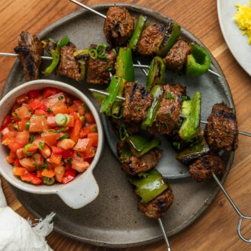 A plate of Mediterranean beef skewers served with a bowl of fresh tomato salsa, turmeric rice just visible to the side.