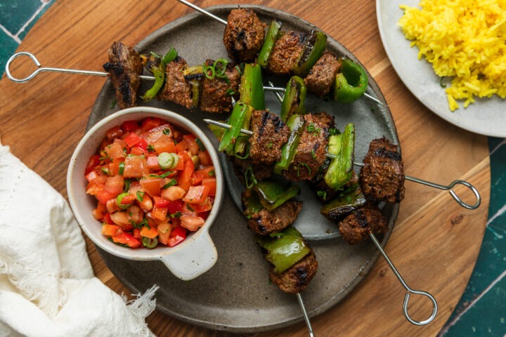 A plate of Mediterranean beef skewers served with a bowl of fresh tomato salsa, turmeric rice just visible to the side.